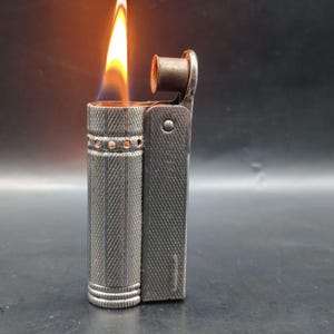 Puede incluir: Un encendedor plateado vintage con una llama encendida. El encendedor es rectangular con un patrón texturizado y bandas decorativas. La llama es amarilla y naranja, con una base azul. El encendedor está abierto, mostrando el mecanismo.