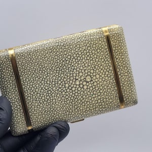 May include: A rectangular case featuring a textured, cream-coloured shagreen surface with a distinct pebbled pattern. The case is accented by two vertical, polished gold-tone metal bands that run across the front, providing a structured contrast.