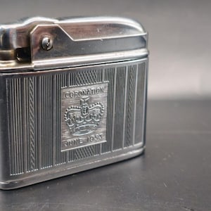 May include: A vintage silver-toned lighter with a rectangular shape. The lighter features vertical line detailing and an engraved emblem with the word "CORONATION" and "JUNE 1953". The lighter has a hinged top and a textured surface.