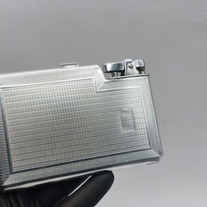 May include: A rectangular, silver-colored cigarette case with a textured, geometric pattern. The case has a hinged lid and a lighter mechanism on the top right corner. The case is held by a gloved hand against a gray background.