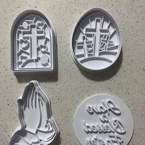 May include: Set of four white plastic cookie cutters featuring religious themes. The cutters are shaped like a cross in a window, three crosses on a hill, praying hands, and a circle with the text "Forever Blessed" in cursive.