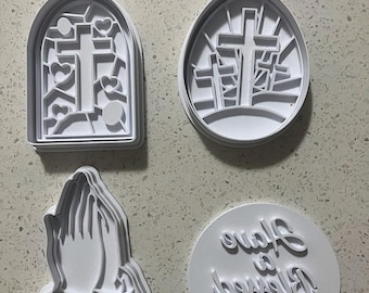 Könnte beinhalten: Satz mit vier weißen Plastik-Ausstechformen mit religiösen Motiven. Die Ausstechformen sind in Form eines Kreuzes in einem Fenster, drei Kreuze auf einem Hügel, betender Hände und eines Kreises mit dem Text "Forever Blessed" in Kursivschrift.