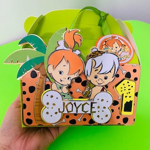 Puede incluir: Una caja de regalo de fiesta verde y naranja con una imagen de dibujos animados de Fred y Wilma Flintstone. La caja tiene un asa y una etiqueta que dice "JOYCE".