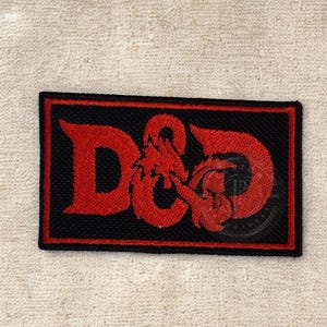 Może przedstawiać: Prostokątna czarna naszywka z czerwonym haftowanym wzorem. Wzór przedstawia litery "D&D" z ilustracją smoka. Naszywka ma czerwoną obwódkę i jest przeznaczona na odzież lub akcesoria.