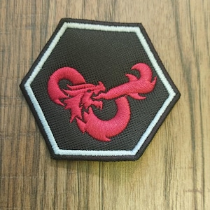 Könnte beinhalten: Ein schwarzer, sechseckiger Aufnäher mit weißem Rand und einem roten Drachendesign. Der Drache ist detailliert gestickt, wobei sein Körper eine stilisierte Form bildet. Der Aufnäher ist wahrscheinlich für Kleidung oder Accessoires.