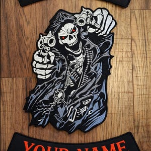 May include: Embroidered patch featuring a grim reaper with red eyes, holding two pistols. The reaper is wearing a black cloak and has a chain around its neck. Two black arched patches with the text "YOUR NAME" in orange are above and below the reaper.