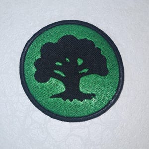 May include: A round patch featuring a black tree silhouette on a green background. The tree is centered within a green circle, which is bordered by a black stitched edge. The patch is likely for clothing or accessories.