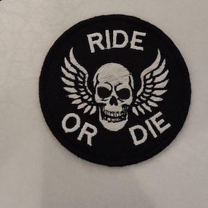 May include: Round black patch with white embroidered text and design. The text reads "RIDE OR DIE" around a white skull with wings. The patch is likely for a jacket or bag.