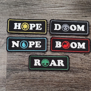以下が含まれることがあります： 白文字とカラフルなアクセントが付いた、5つの長方形の黒いパッチ。「HOPE」、「DOOM」、「NOPE」、「BOOM」、「ROAR」と書かれています。各パッチには、太陽、スカル、水滴、火、木など、異なるシンボルが付いています。