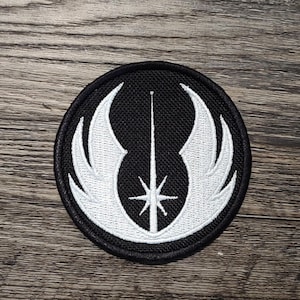 May include: A round, black patch with a white emblem featuring a central vertical line, a star, and stylized wings. The patch is embroidered with a detailed design, suitable for application on clothing or accessories.