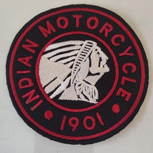 Może przedstawiać: Okrągła czarna naszywka z czerwoną obwódką i napisem "INDIAN MOTORCYCLE 1901". Naszywka przedstawia biały profil indiańskiego wodza. Tekst i wzór są haftowane.