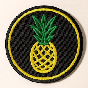 May include: Round black patch with a yellow border and a yellow pineapple design in the center. The pineapple has a green leafy top. The patch is likely an iron-on or sew-on patch for clothing or accessories.
