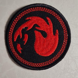 May include: Round black patch with a red flame design. The flame is in the center, with a detailed red border and stitching. The patch is likely for clothing or accessories.