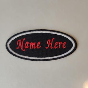 May include: An oval black patch with the embroidered text "Name Here" in red script. The patch has a white border. This patch is designed for customisation, likely for clothing or accessories, offering a personalized touch.