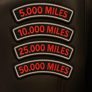 May include: Close-up of a black leather jacket with four embroidered patches. Each patch has a white border and red text indicating mileage milestones: 5,000, 10,000, 25,000, and 50,000 miles. The jacket has visible stitching.
