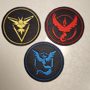 Puede incluir: Tres parches redondos bordados con fondos negros. Uno presenta un diseño de rayo amarillo, otro un pájaro rojo y el tercero una criatura azul con forma de pájaro. Cada parche tiene un borde negro.