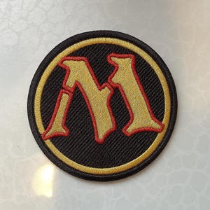 Könnte beinhalten: Runder, gestickter Aufnäher mit einem goldenen Buchstaben "M" und roten Akzenten auf schwarzem Hintergrund. Das Design wird von einem goldenen Rand umrahmt. Geeignet für Kleidung oder Accessoires, dient dieser Aufnäher dekorativen Zwecken.