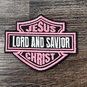 May include: A black and pink embroidered patch with the words "JESUS LORD AND SAVIOR CHRIST" in white and pink lettering. The patch has a shield shape with a black border.