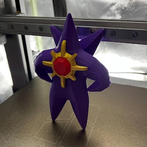 May include: A 3D-printed purple star-shaped figure with a gold and red center. The figure has a pointed top and five arms. The object is standing on a textured surface.