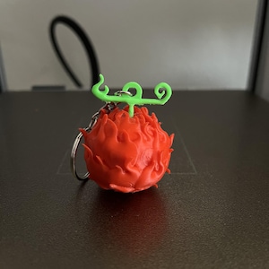 Op de afbeelding: Een rode, getextureerde sleutelhanger in de vorm van een vrucht met een groene, gekrulde stengel. De sleutelhanger is bevestigd aan een zilveren ring. De vrucht heeft een gedetailleerd, hobbelig oppervlak.