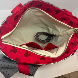 Puede incluir: Bolso tote abierto con interior rojo con siluetas negras de Mickey Mouse. El bolso tiene un forro color crema, un cierre de cremallera y un bolsillo peque&ntilde;o. El exterior presenta un estampado de cuadros grises y blancos y un lazo gris con lunares negros.