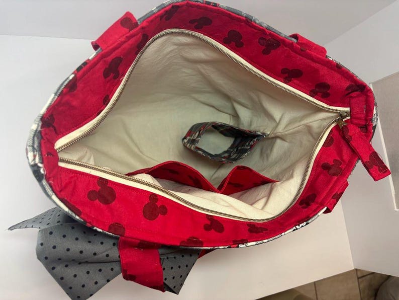 Puede incluir: Bolso tote abierto con interior rojo con siluetas negras de Mickey Mouse. El bolso tiene un forro color crema, un cierre de cremallera y un bolsillo peque&ntilde;o. El exterior presenta un estampado de cuadros grises y blancos y un lazo gris con lunares negros.