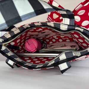 May include: A black and white checkered tote bag with a red polka dot bow and interior lining. The bag is open, revealing a pink tumbler, a wallet, and a laptop. The bag has a zipper closure and a black and white checkered strap.