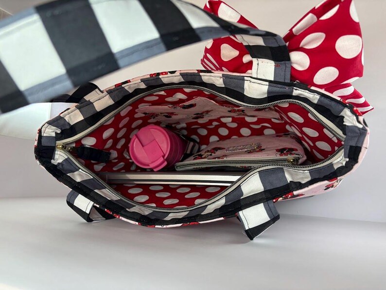 May include: A black and white checkered tote bag with a red polka dot bow and interior lining. The bag is open, revealing a pink tumbler, a wallet, and a laptop. The bag has a zipper closure and a black and white checkered strap.