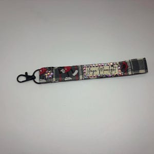 Puede incluir: Un llavero de pulsera de tela con un cierre negro. El llavero presenta un estampado de cuadros en gris, rojo y negro. El texto "Yaya Moonlight" se muestra en el llavero, rodeado de pedrer&iacute;a.