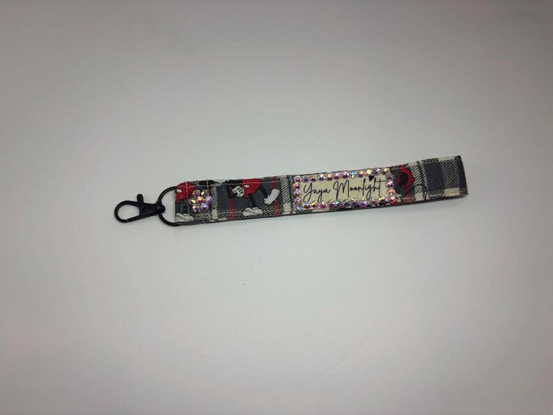 Puede incluir: Un llavero de pulsera de tela con un cierre negro. El llavero presenta un estampado de cuadros en gris, rojo y negro. El texto "Yaya Moonlight" se muestra en el llavero, rodeado de pedrer&iacute;a.