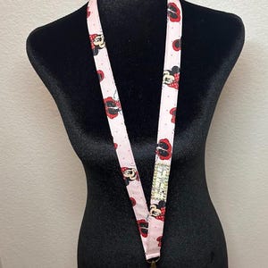 May include: Pink lanyard featuring a repeating pattern of Minnie Mouse with red bows and polka dots. The lanyard is displayed on a black mannequin and has a metal clasp at the bottom. A small tag with text is visible.