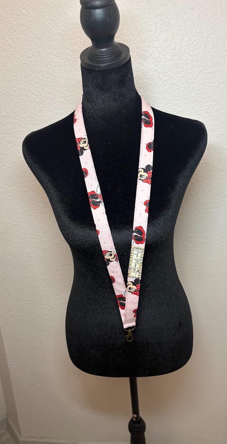 May include: Pink lanyard featuring a repeating pattern of Minnie Mouse with red bows and polka dots. The lanyard is displayed on a black mannequin and has a metal clasp at the bottom. A small tag with text is visible.