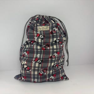 Puede incluir: Una bolsa con cord&oacute;n de cuadros grises con un estampado de Mickey y Minnie Mouse. La bolsa tiene un cierre de cord&oacute;n gris y una peque&ntilde;a etiqueta rectangular. La bolsa est&aacute; hecha de tela y es adecuada para transportar art&iacute;culos.