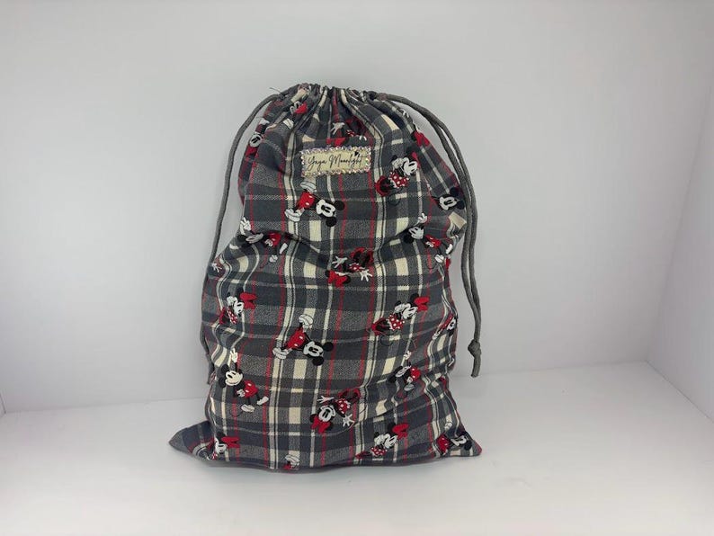 Puede incluir: Una bolsa con cord&oacute;n de cuadros grises con un estampado de Mickey y Minnie Mouse. La bolsa tiene un cierre de cord&oacute;n gris y una peque&ntilde;a etiqueta rectangular. La bolsa est&aacute; hecha de tela y es adecuada para transportar art&iacute;culos.