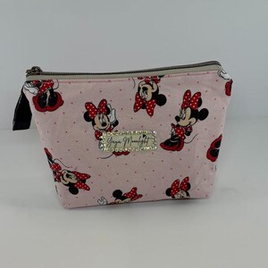 May include: A pink zippered cosmetic bag featuring a pattern of Minnie Mouse in various poses with red bows. The bag has a black zipper pull and a rectangular rhinestone embellishment with the text "Yaya Moonlight".
