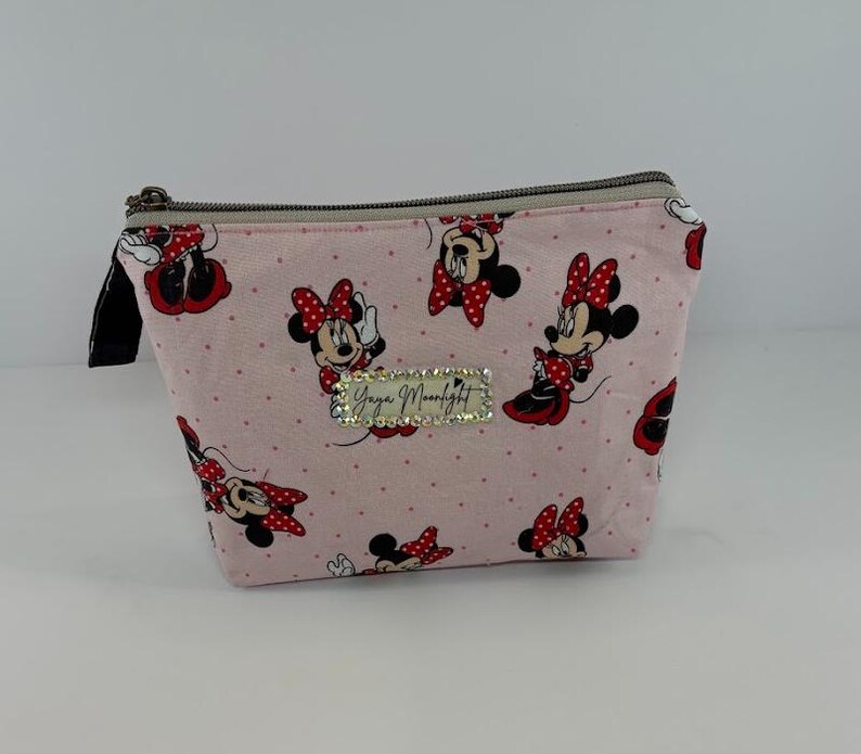 May include: A pink zippered cosmetic bag featuring a pattern of Minnie Mouse in various poses with red bows. The bag has a black zipper pull and a rectangular rhinestone embellishment with the text "Yaya Moonlight".