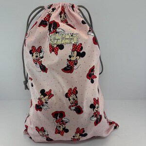 May include: A pink drawstring bag featuring a repeating pattern of Minnie Mouse in various poses, with red bows and polka dots. The bag has a gray drawstring closure and a small rectangular label with text.
