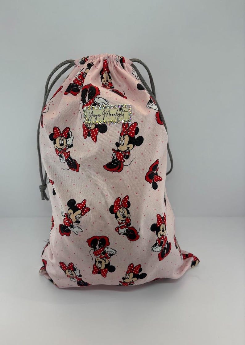 May include: A pink drawstring bag featuring a repeating pattern of Minnie Mouse in various poses, with red bows and polka dots. The bag has a gray drawstring closure and a small rectangular label with text.