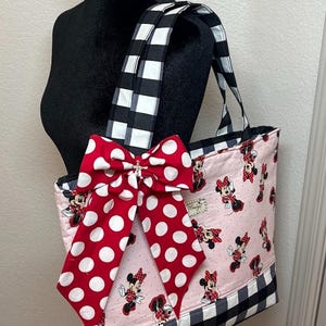 May include: Pink tote bag featuring a Minnie Mouse pattern, a large red bow with white polka dots, and black and white checkered accents. The bag has matching checkered handles and a black base.