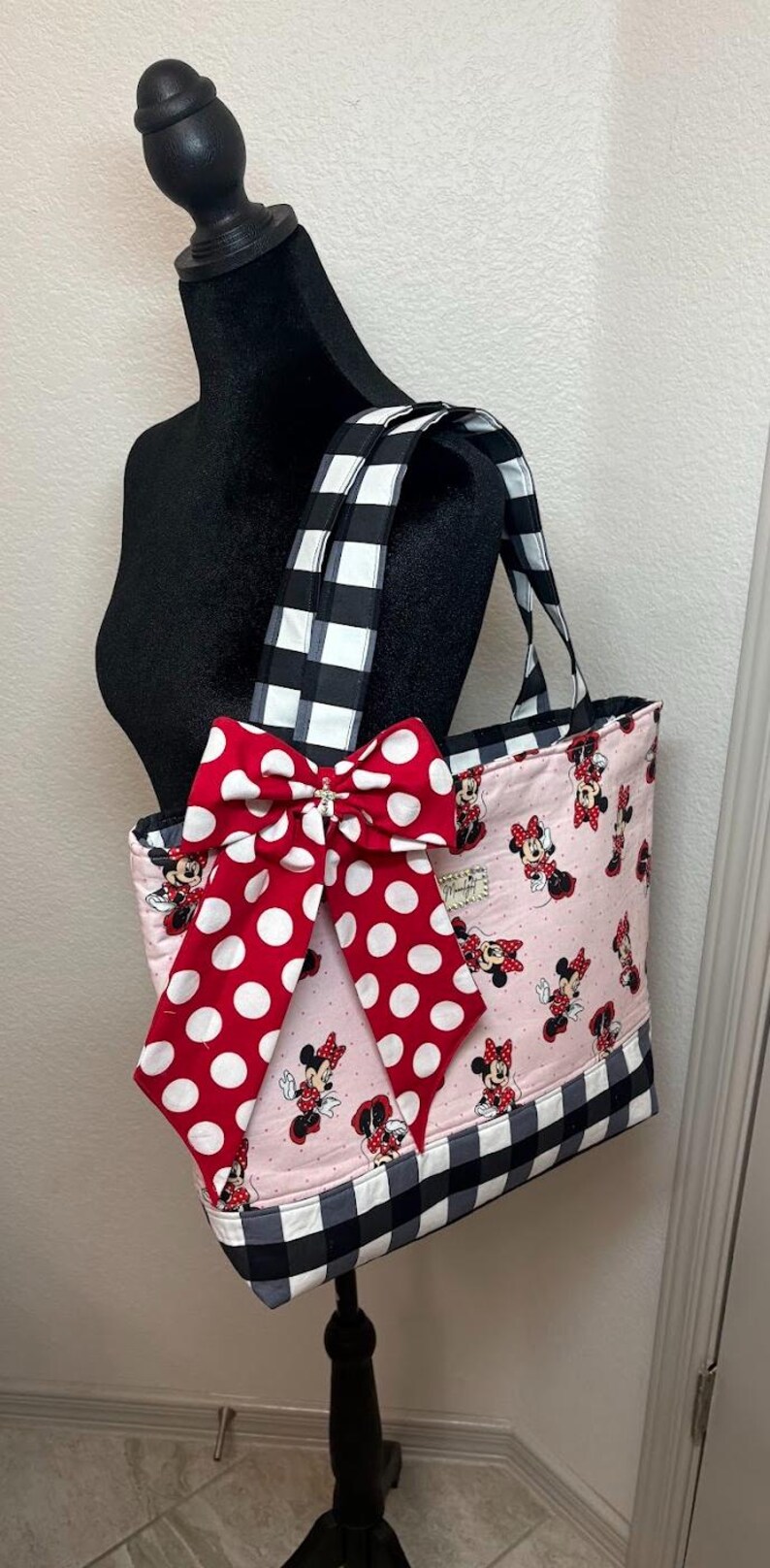 May include: Pink tote bag featuring a Minnie Mouse pattern, a large red bow with white polka dots, and black and white checkered accents. The bag has matching checkered handles and a black base.
