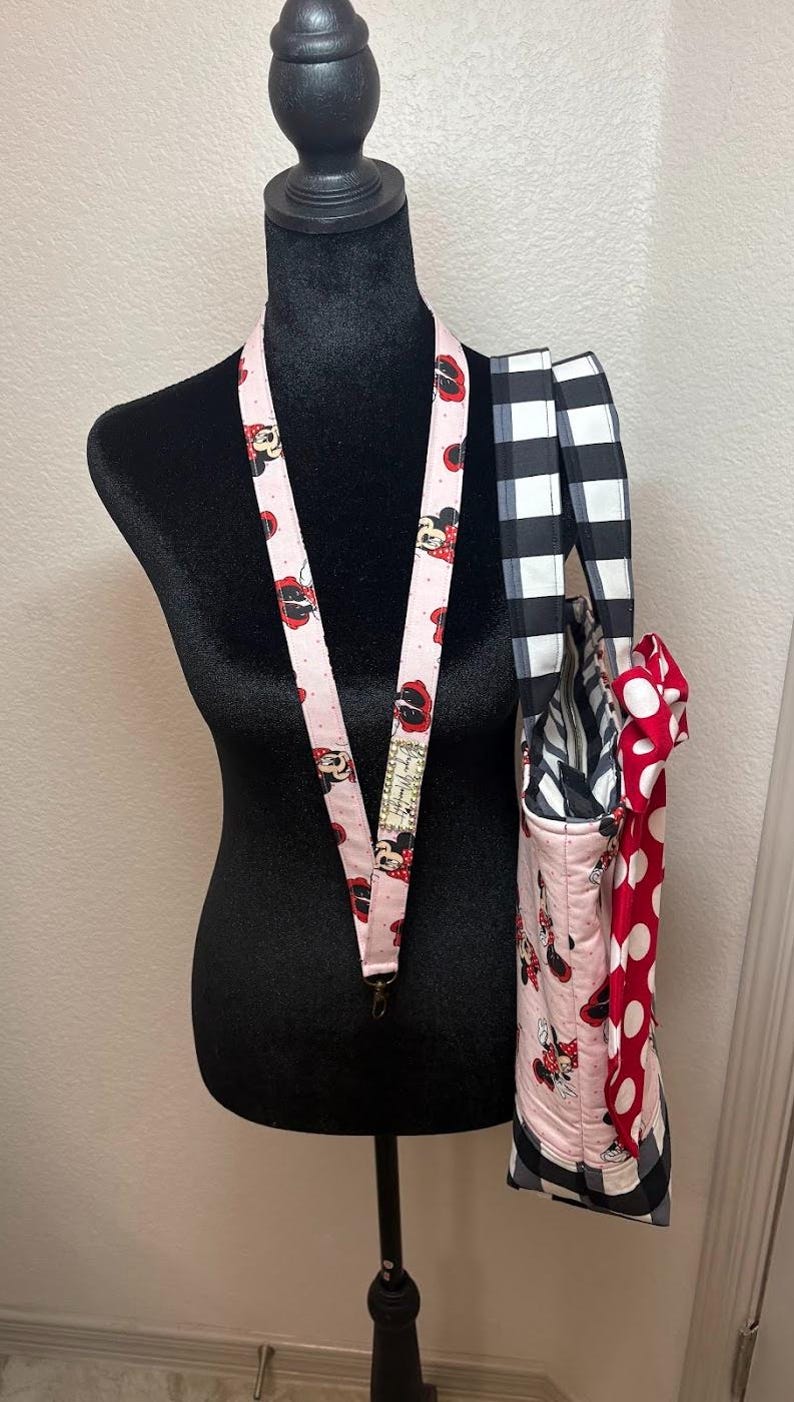 May include: A pink lanyard with a Minnie Mouse pattern hangs on a black mannequin. A matching tote bag with a black and white checkered strap and a red polka dot bow is also displayed. The tote bag has a Minnie Mouse pattern.