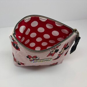 May include: Pink zippered cosmetic bag with a Minnie Mouse pattern. The bag features a red interior with white polka dots. The exterior has a repeating pattern of Minnie Mouse figures. A black pull tab is attached to the zipper.