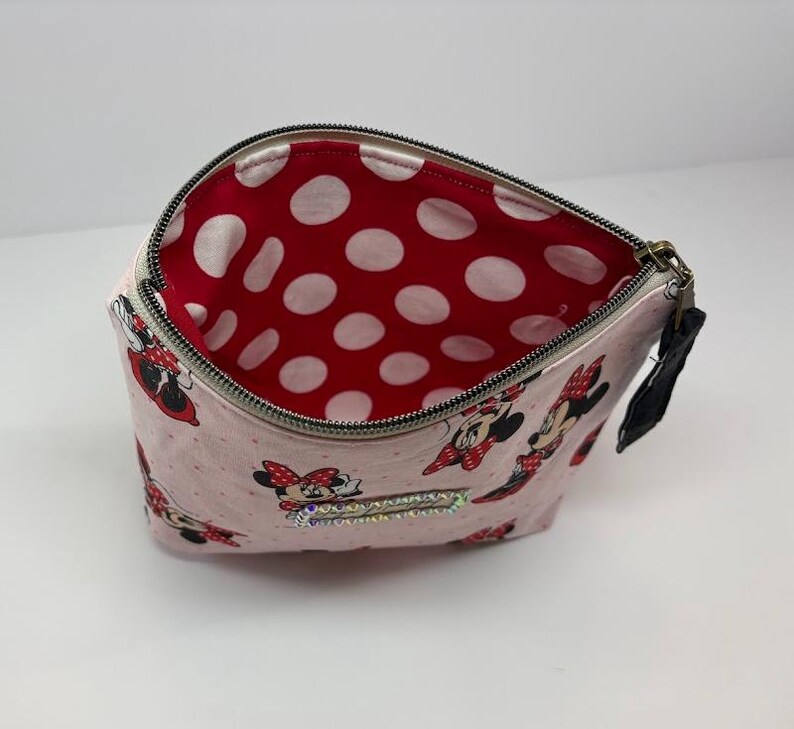 May include: Pink zippered cosmetic bag with a Minnie Mouse pattern. The bag features a red interior with white polka dots. The exterior has a repeating pattern of Minnie Mouse figures. A black pull tab is attached to the zipper.