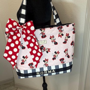 May include: A pink tote bag with Minnie Mouse illustrations, a large red polka dot bow, and black and white checkered details. The name "NORMA" is displayed on the front. The bag has black and white checkered handles.