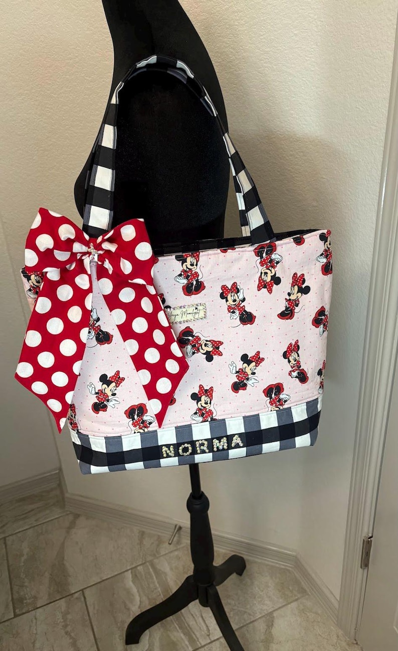 May include: A pink tote bag with Minnie Mouse illustrations, a large red polka dot bow, and black and white checkered details. The name "NORMA" is displayed on the front. The bag has black and white checkered handles.