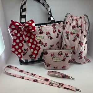 May include: A coordinated set of accessories with a Minnie Mouse design. The set includes a tote bag with a black and white check pattern, a drawstring bag, a cosmetic bag, a lanyard, and a wristlet. All items are pink with a Minnie Mouse print and a red polka dot bow.