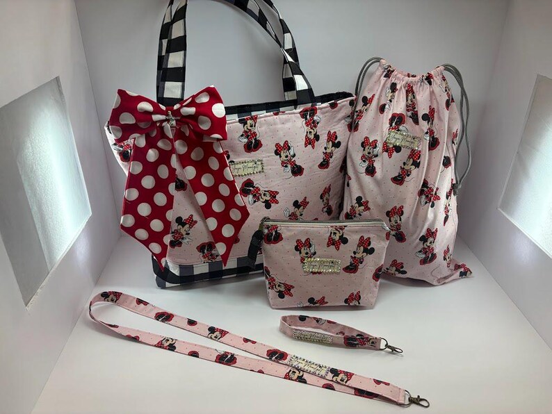 May include: A coordinated set of accessories with a Minnie Mouse design. The set includes a tote bag with a black and white check pattern, a drawstring bag, a cosmetic bag, a lanyard, and a wristlet. All items are pink with a Minnie Mouse print and a red polka dot bow.