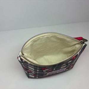 Puede incluir: Bolsa de cosm&eacute;ticos abierta con un estampado de cuadros y detalles en rojo y blanco. El interior es beige. La bolsa tiene un cierre de cremallera con un tirador rojo. La bolsa es de tela.