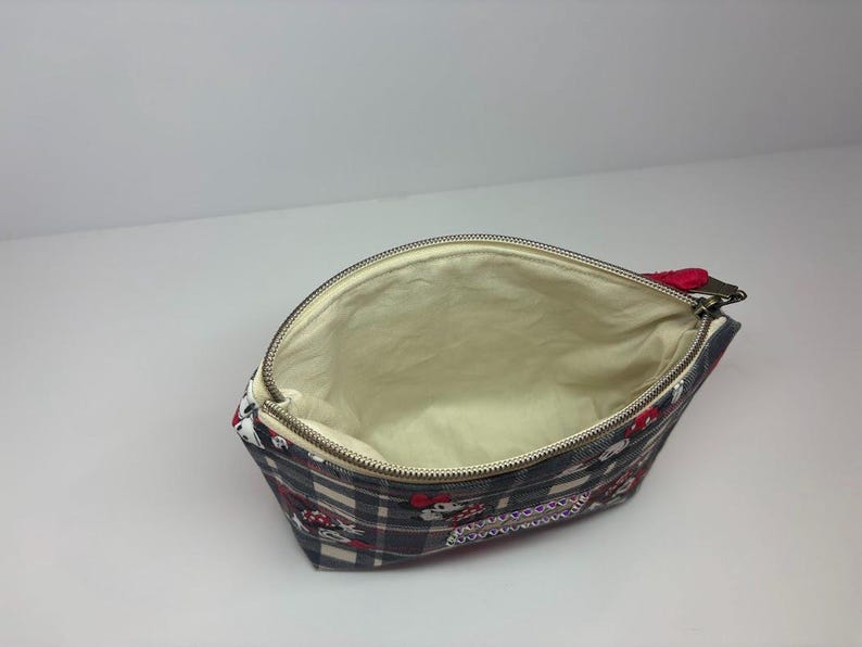 Puede incluir: Bolsa de cosm&eacute;ticos abierta con un estampado de cuadros y detalles en rojo y blanco. El interior es beige. La bolsa tiene un cierre de cremallera con un tirador rojo. La bolsa es de tela.