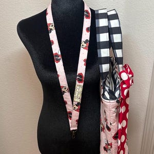 May include: A pink Minnie Mouse lanyard with a metal clasp hangs on a black mannequin. A matching tote bag with a black and white checked strap and a red polka dot bow is also displayed.